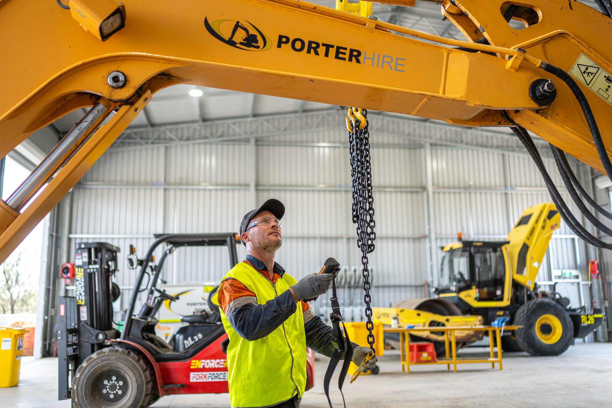 A Porter Hire worker in high visibility clothing inspects a chain hanging from a yellow excavator arm inside a machinery workshop. Forklifts and heavy equipment in the background show a busy equipment hire and maintenance setting.