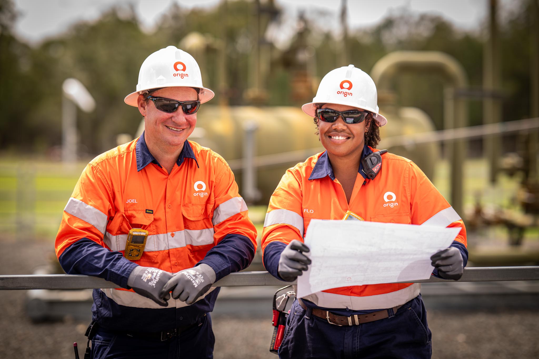 Two Origin Energy workers smile while standing at an industrial site in hard hats, sunglasses and high visibility uniforms. One worker holds site plans with pipelines and equipment visible in the background.