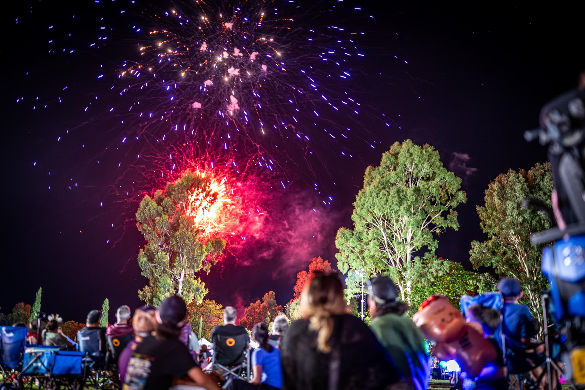 A crowd watches red and blue fireworks burst above trees during a night time outdoor event. People sit in camping chairs and look toward the bright display, creating a relaxed community festival atmosphere.
