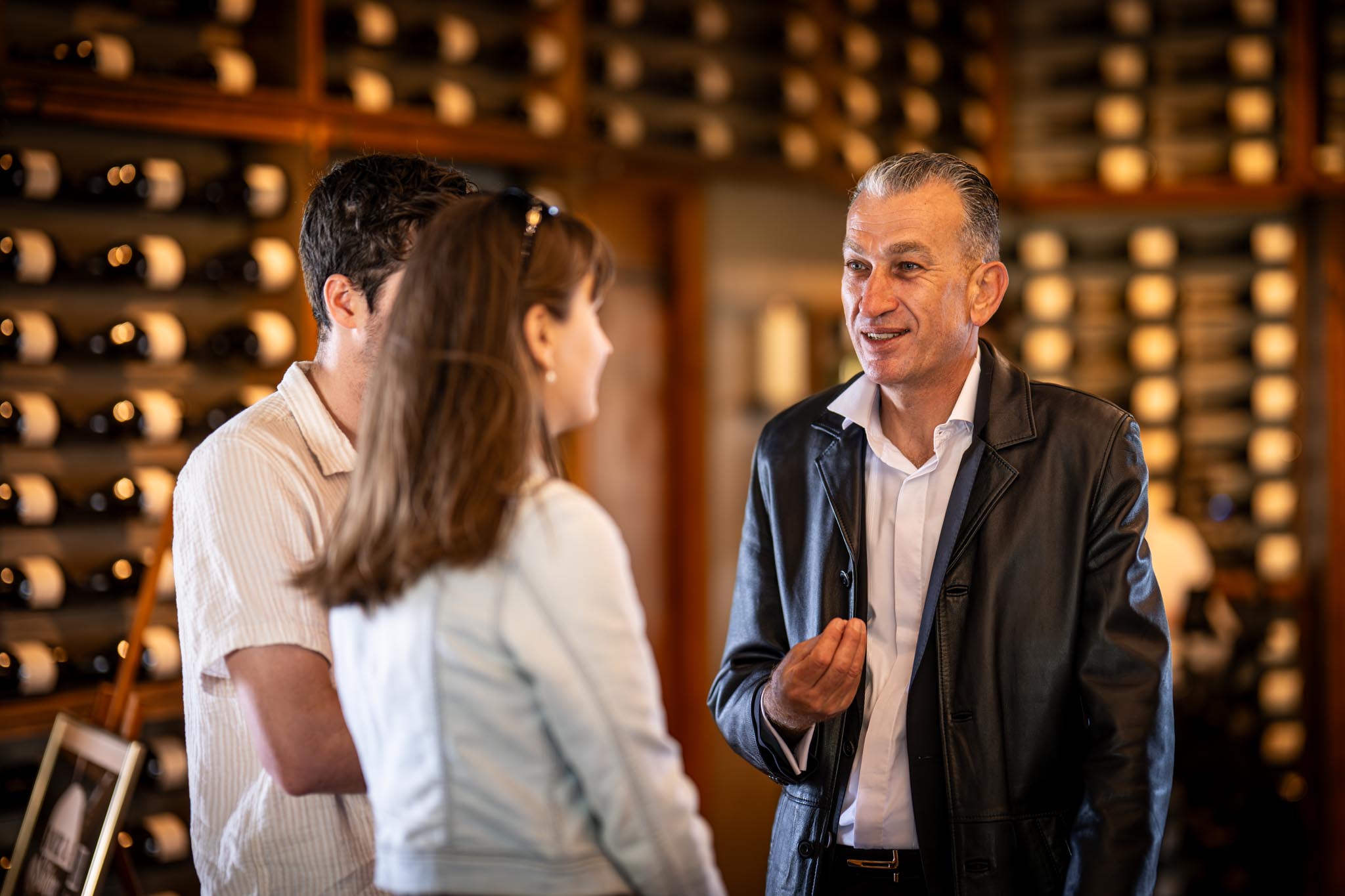 A man in a leather jacket speaks with two guests inside a warmly lit wine cellar lined with bottles. The setting suggests a guided tasting or hospitality experience in a premium venue.