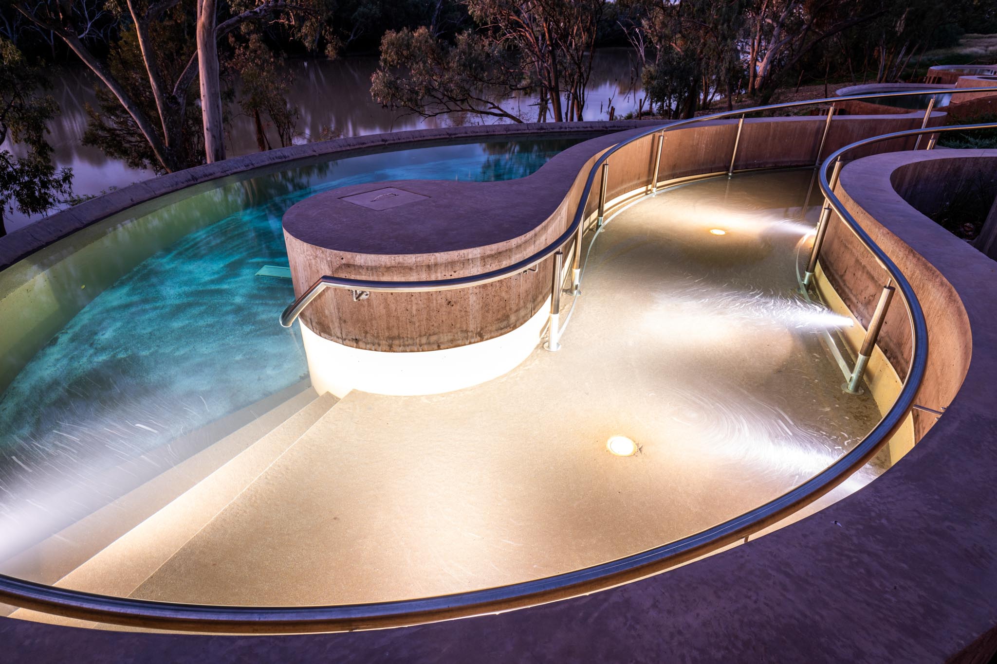 Curved Cunnamulla Hot Springs pools glow with underwater lighting beside trees and a river at dusk. Stainless steel handrails, concrete edges and warm water jets create a calm outdoor bathing area.