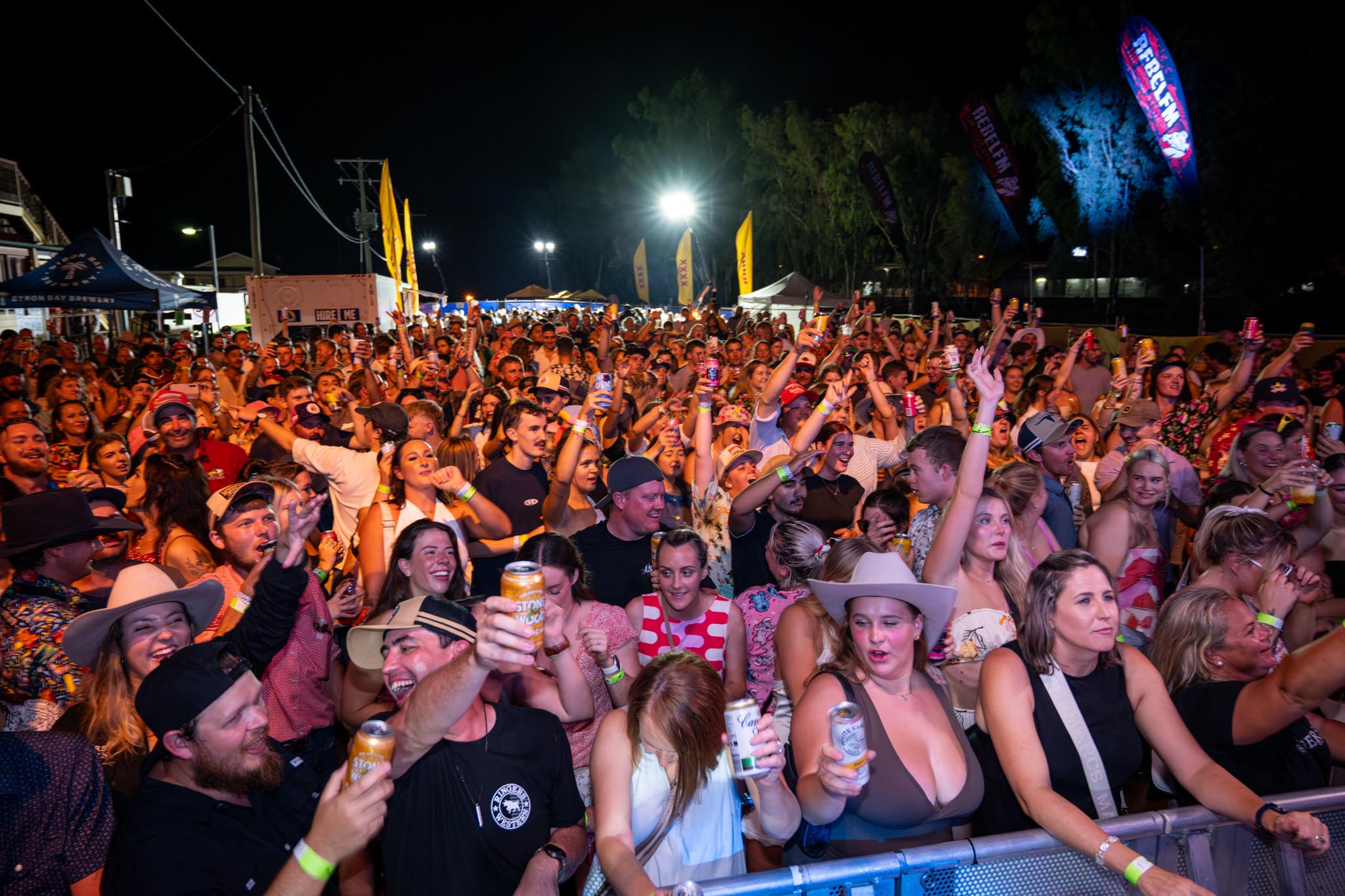A large crowd at Chinchilla Melon Festival cheers with raised drinks during a night time live event. Bright stage lighting, festival flags and packed front row barriers show an energetic concert atmosphere.