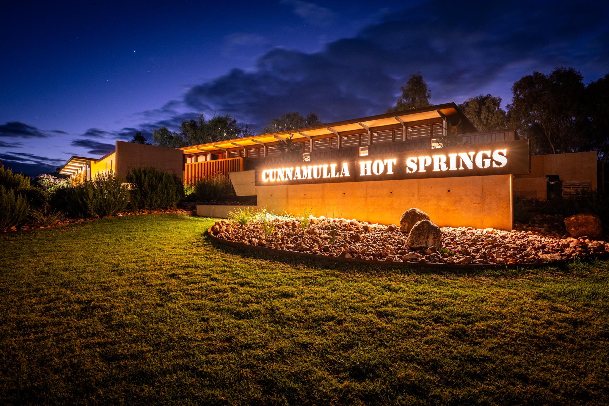 Illuminated entrance sign reading CUNNAMULLA HOT SPRINGS glows in front of the hot springs building at night. Warm lighting, landscaped rocks and a deep blue sky create a welcoming resort style arrival scene.