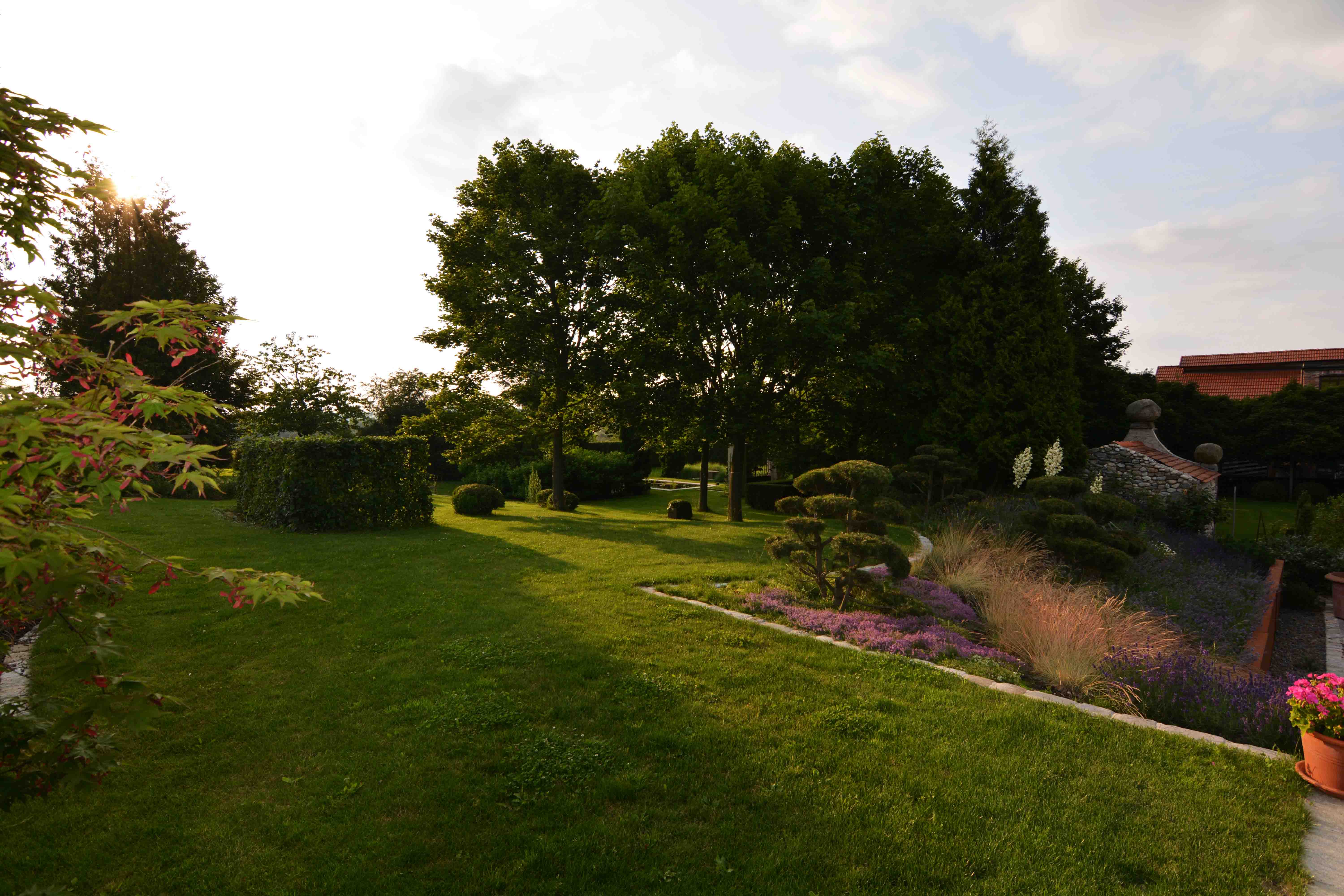 Grüner Garten mit gepflegtem Rasen, Büschen, Bäumen, Blumenbeeten und einem Steinmauerabschnitt bei Sonnenuntergang.