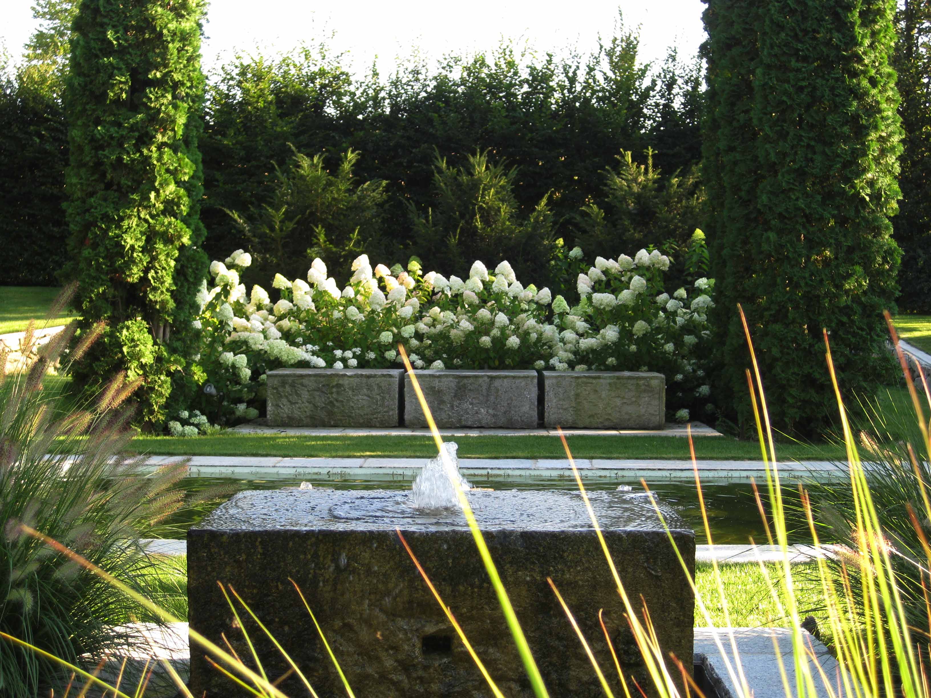 Steinerner Wasserspender mit Springbrunnen vor weißen Blumenbüschen und großen grünen Bäumen in einem gepflegten Garten.