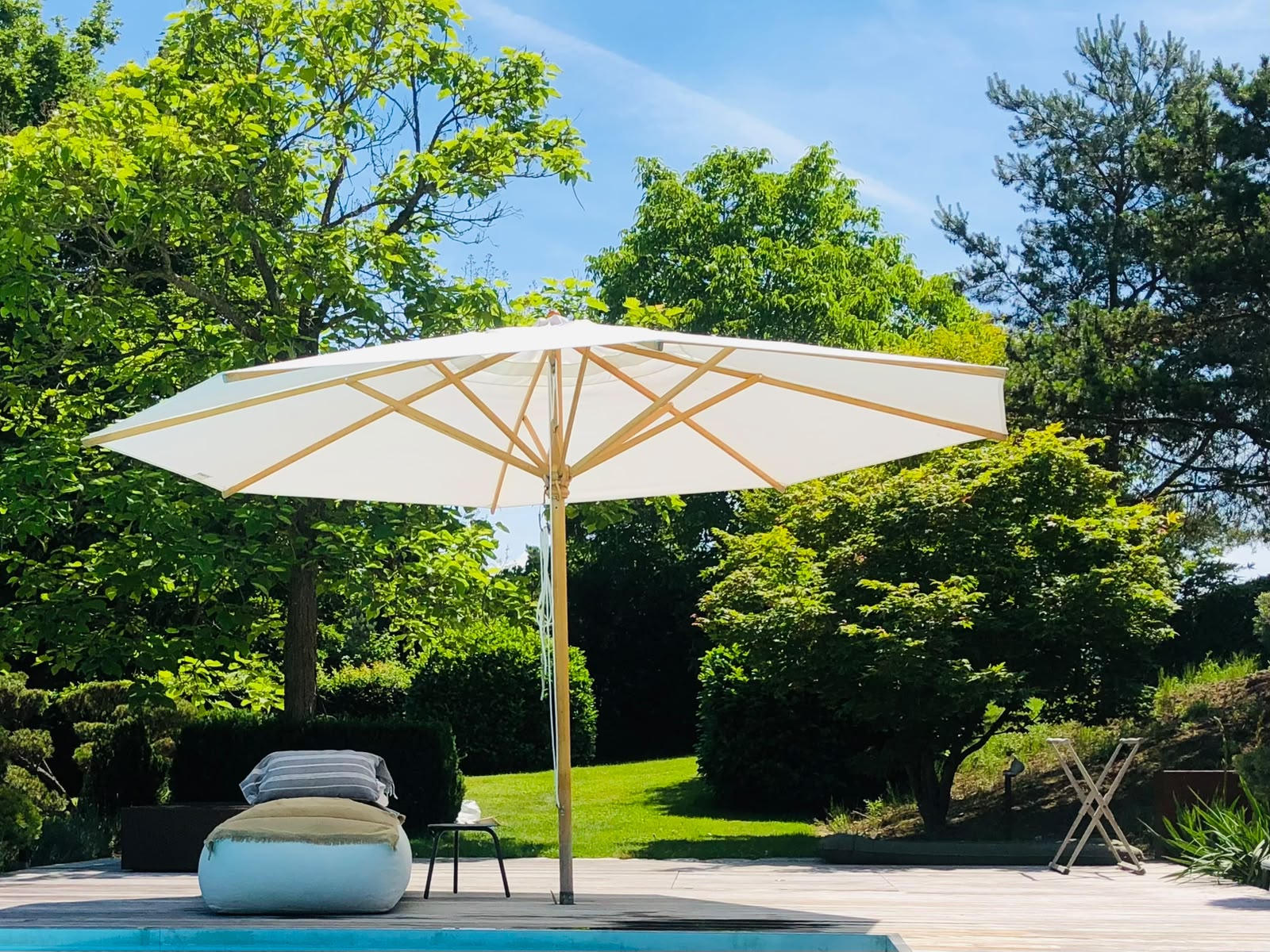 Weißer Sonnenschirm und Liegestuhl neben Pool auf Holzterrasse umgeben von grünen Bäumen.