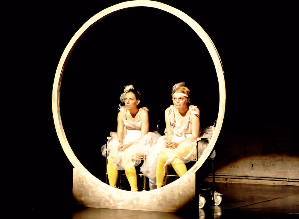 Zwei Schauspielerinnen, die nebeneinander auf der Bühne sitzen, blicken durch einen großen Holzreifen