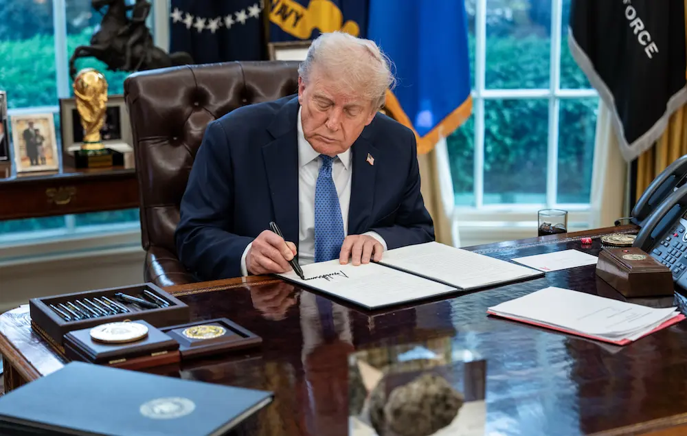 Man in dark suit signing a large book at a polished desk in an office with a window and flags behind him.