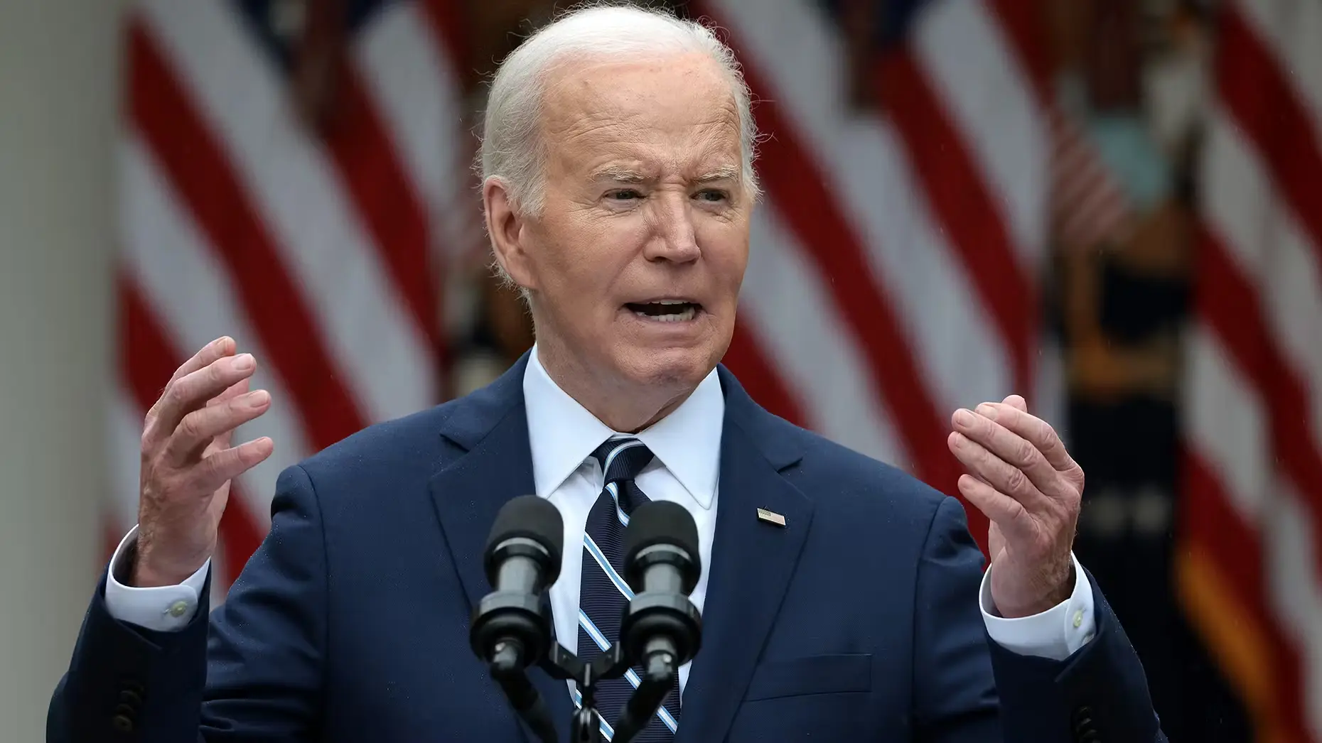 Man in navy suit speaking at podium with blurred American flags in the background.