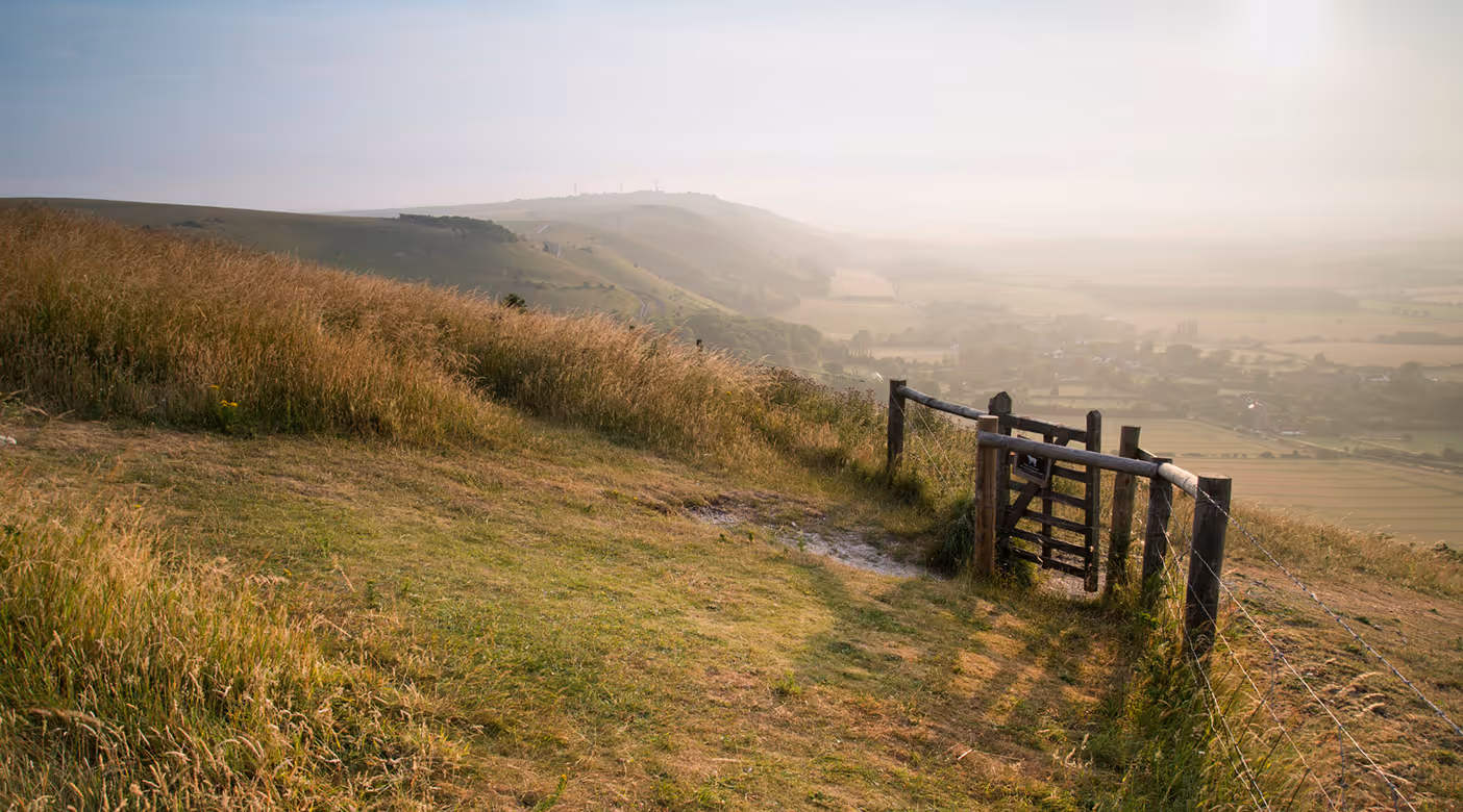 Countryside landscape with a grassy hill, a wooden gate fence, and a misty valley in the background.
