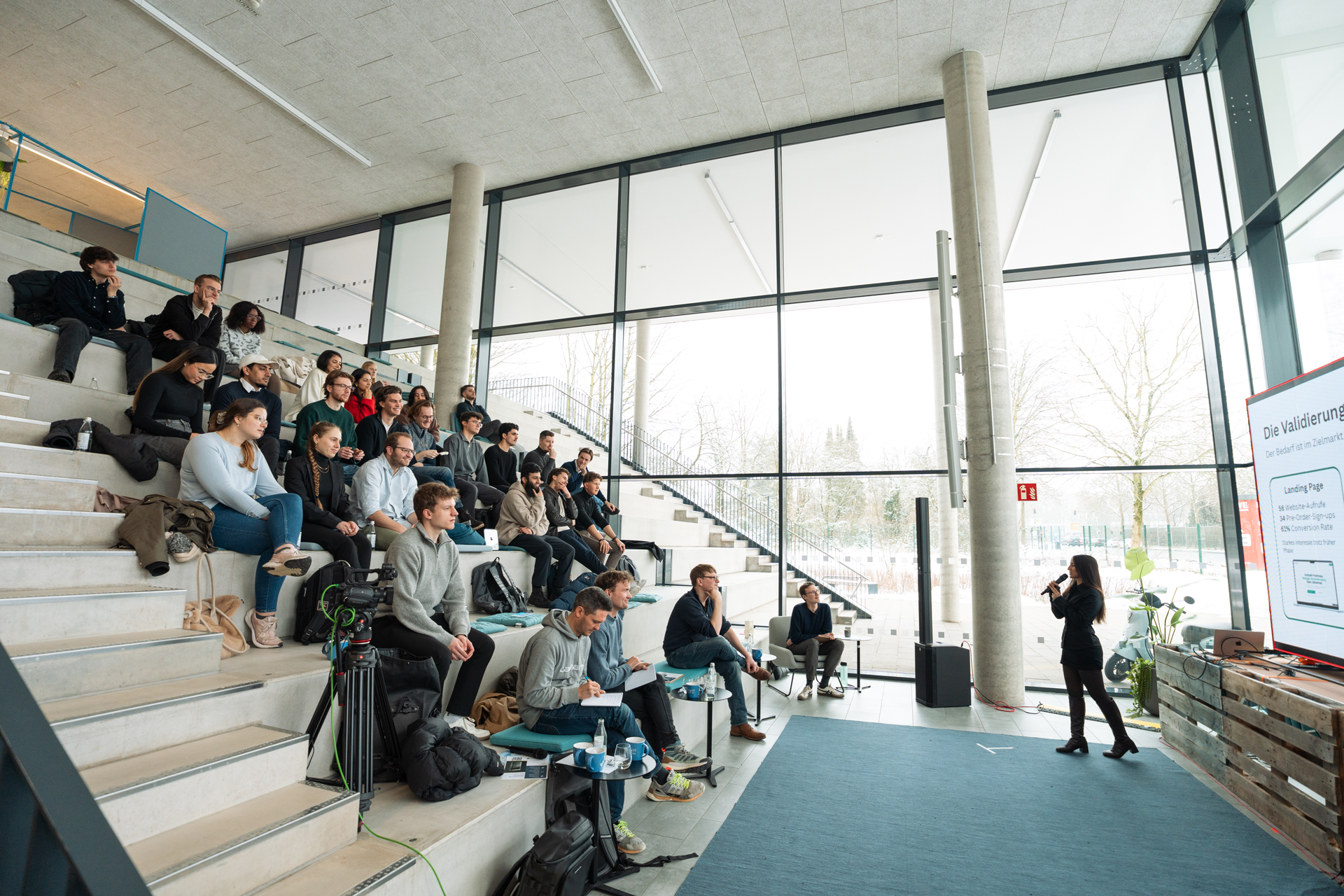 Speaker presenting to a seated audience in a modern room with large windows and tiered concrete seating.