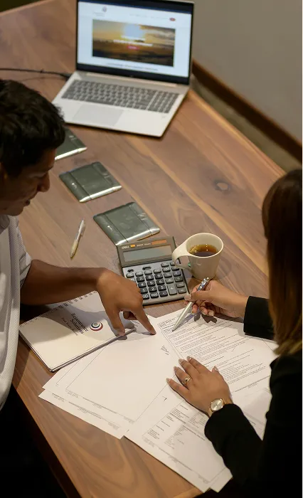 Two colleagues reviewing documents at a desk with a laptop and calculator