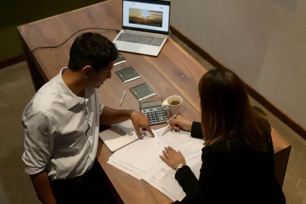 Two colleagues reviewing documents at a desk with a laptop and calculator