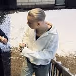 Person med långt blont hår samlat i en knut, iförd en vit tröja och gröna byxor, stirrar nedåt medan de står på en trappa med snö och en annan persons hand syns till vänster.