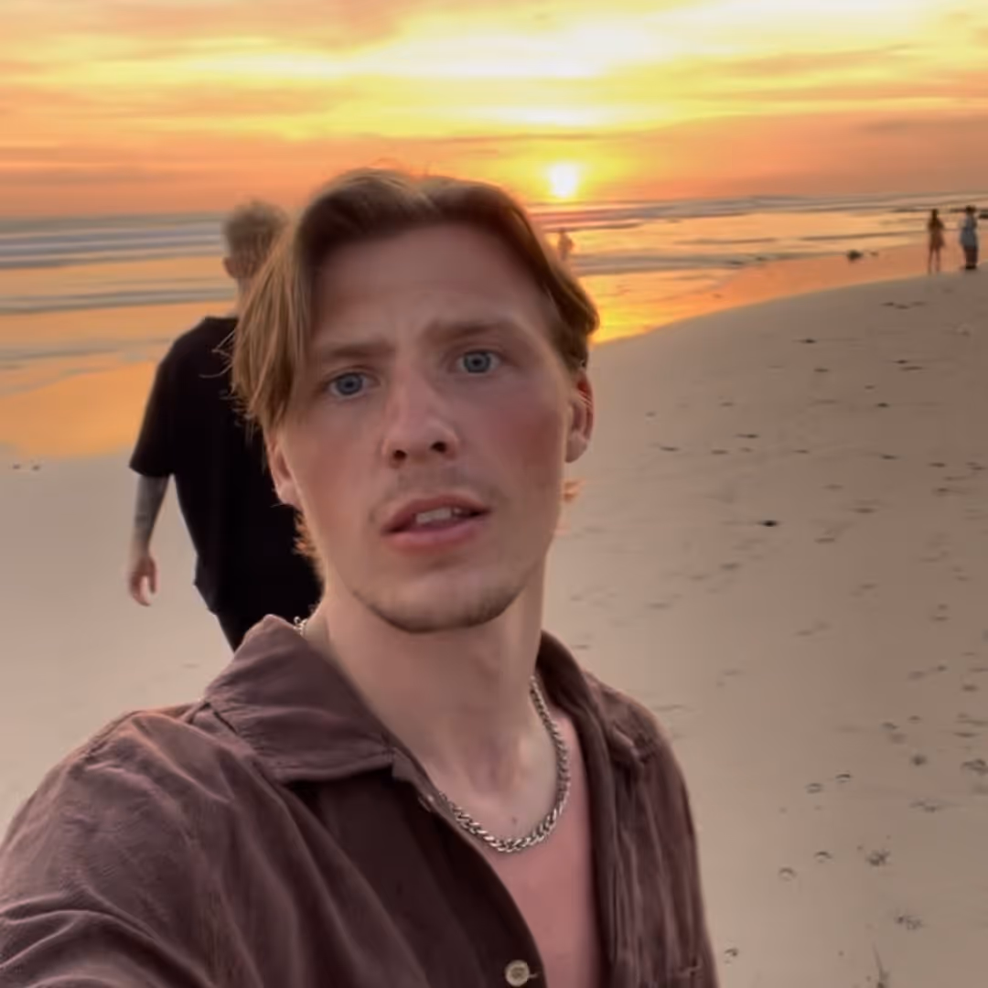 Ung man med brunt skjorta och kedjehalsband tar en selfie på en strand under solnedgången.