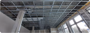 Decke in einem Raum während Renovierungsarbeiten mit sichtbarem Metalluntergerüst und großen Fenstern an der rechten Wand.