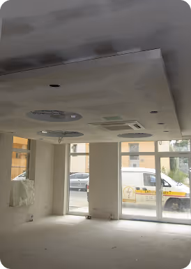 Innenraum eines unfertigen, leeren Raumes mit eingebauter Deckenbeleuchtung und großen Fenstern, durch die ein geparkter Lieferwagen zu sehen ist.