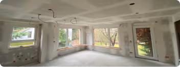 Unfertiges Zimmer mit großen Fenstern, weißen Wänden mit Gipsarbeiten und sichtbaren Stromleitungen.