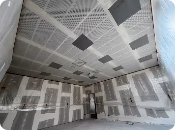 Rohbau eines Raums mit teilweise verkleideten Wänden und dunklen rechteckigen Akustikplatten an der Decke.