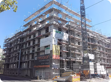 Mehrstöckiges Gebäude im Rohbau mit Gerüsten und Baukran an einer sonnigen Straße.