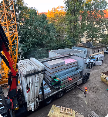 Der Lastwagen auf einer Baustelle ist mit Bauholz, Dämmmaterialien und anderen Baumaterialien beladen, umgeben von Bäumen und Gebäuden im Hintergrund.