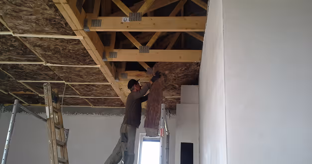 Mann installiert Dämmmaterial an einer Holzdecke in einem Raum mit weißen Wänden.