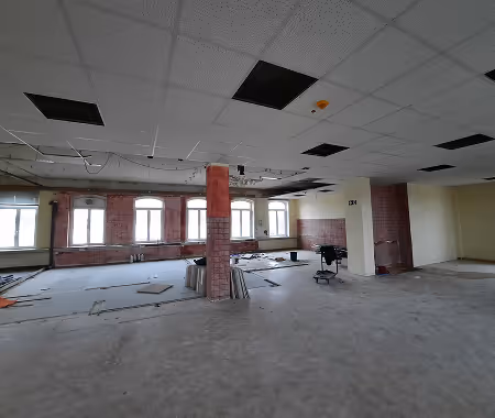 Großer leerer Raum mit unvollständiger Decke und freiliegenden Ziegelwänden, Baustellenmaterialien und Stuhl auf Boden.