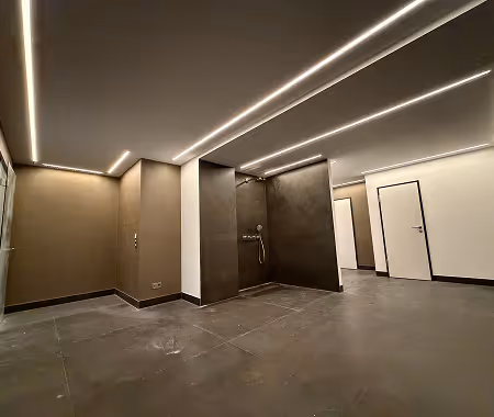 Moderne, minimalistische Umkleide mit braunen Wänden, beleuchteter Decke und Duschbereich.