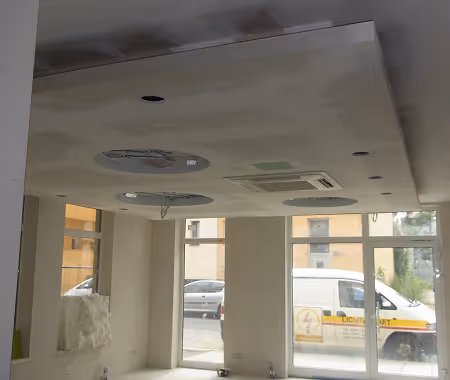 Innenraum mit unvollständig montierter abgehängter Decke mit runden Aussparungen, großen Fenstern und einem weißen Lieferwagen draußen.