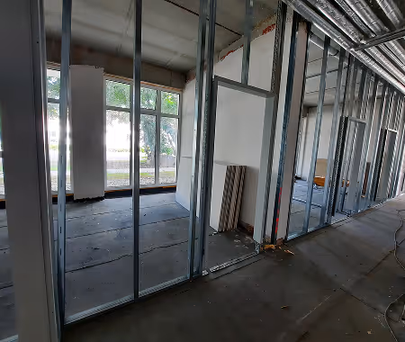 Innenansicht einer Baustelle mit Metallrahmen für Wände und großen Fenstern in einem Raum mit Betonboden.