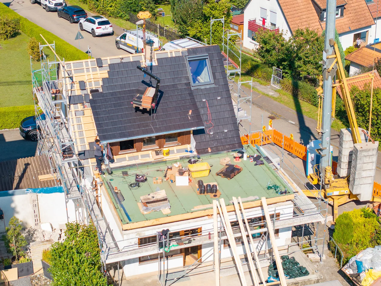 Vogelperspektive einer Baustelle zur Dachsanierung an einem Wohnhaus mit Gerüst, Baukran und frisch verlegten dunklen Dachziegeln.