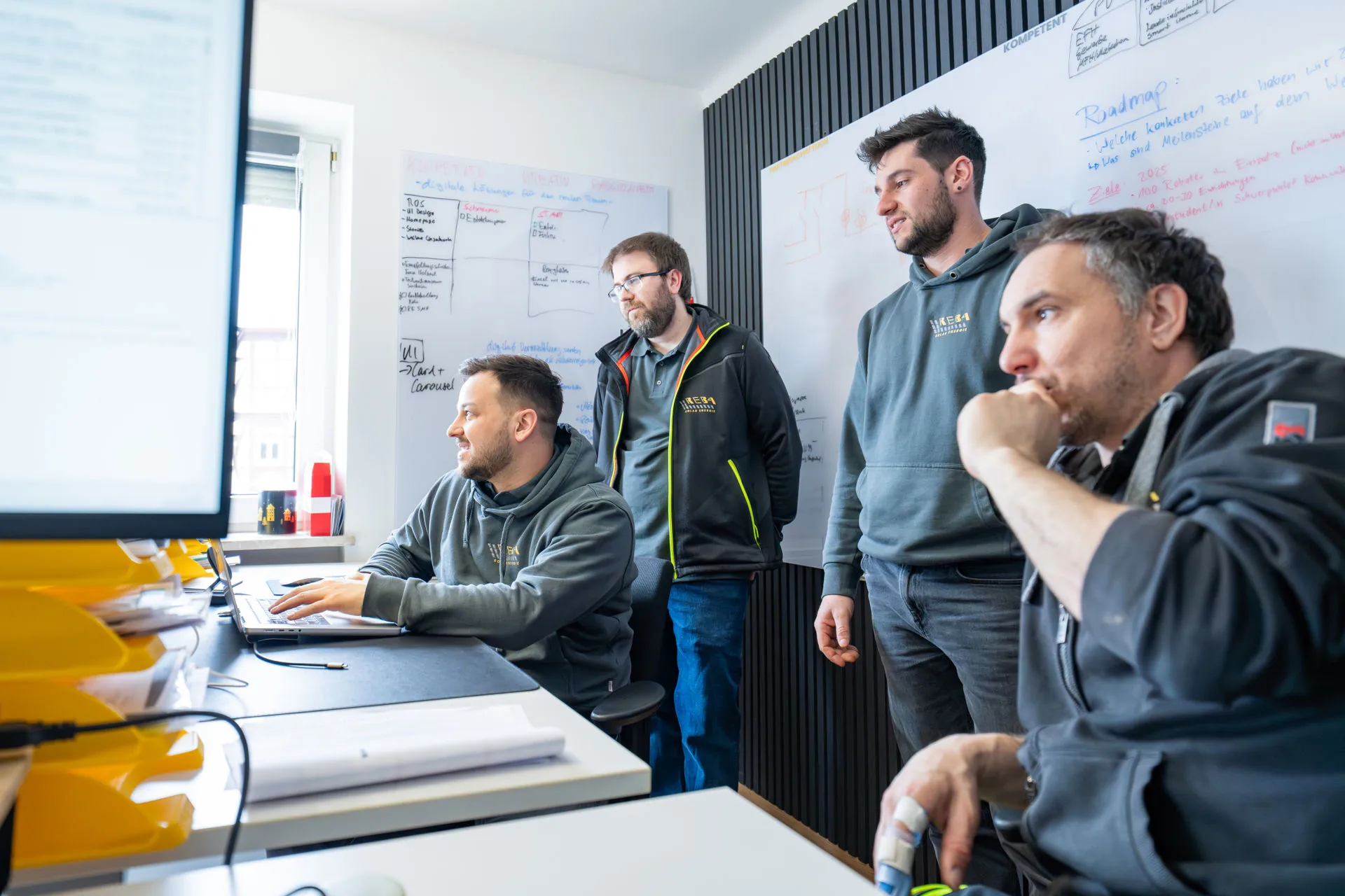 Ein Team von Reba Solar bei der Projektplanung im Büro; vier Mitarbeiter besprechen Details an einem Computerbildschirm vor einem Whiteboard mit einer Roadmap.