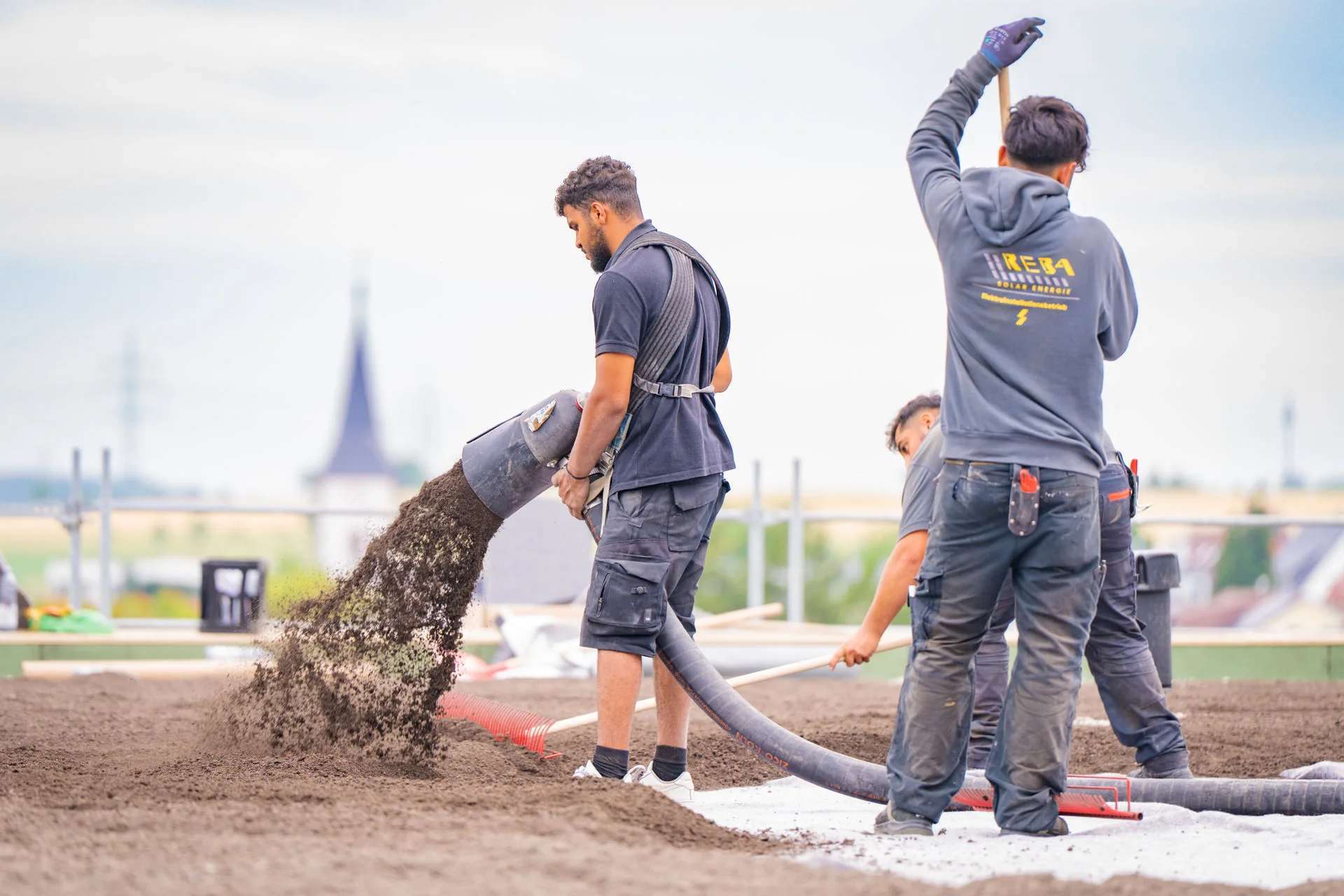 Drei Handwerker von Reba Solar bringen mit einem Schlauch Substrat für eine Dachbegrünung auf einem Flachdach aus und verteilen es mit Rechen.