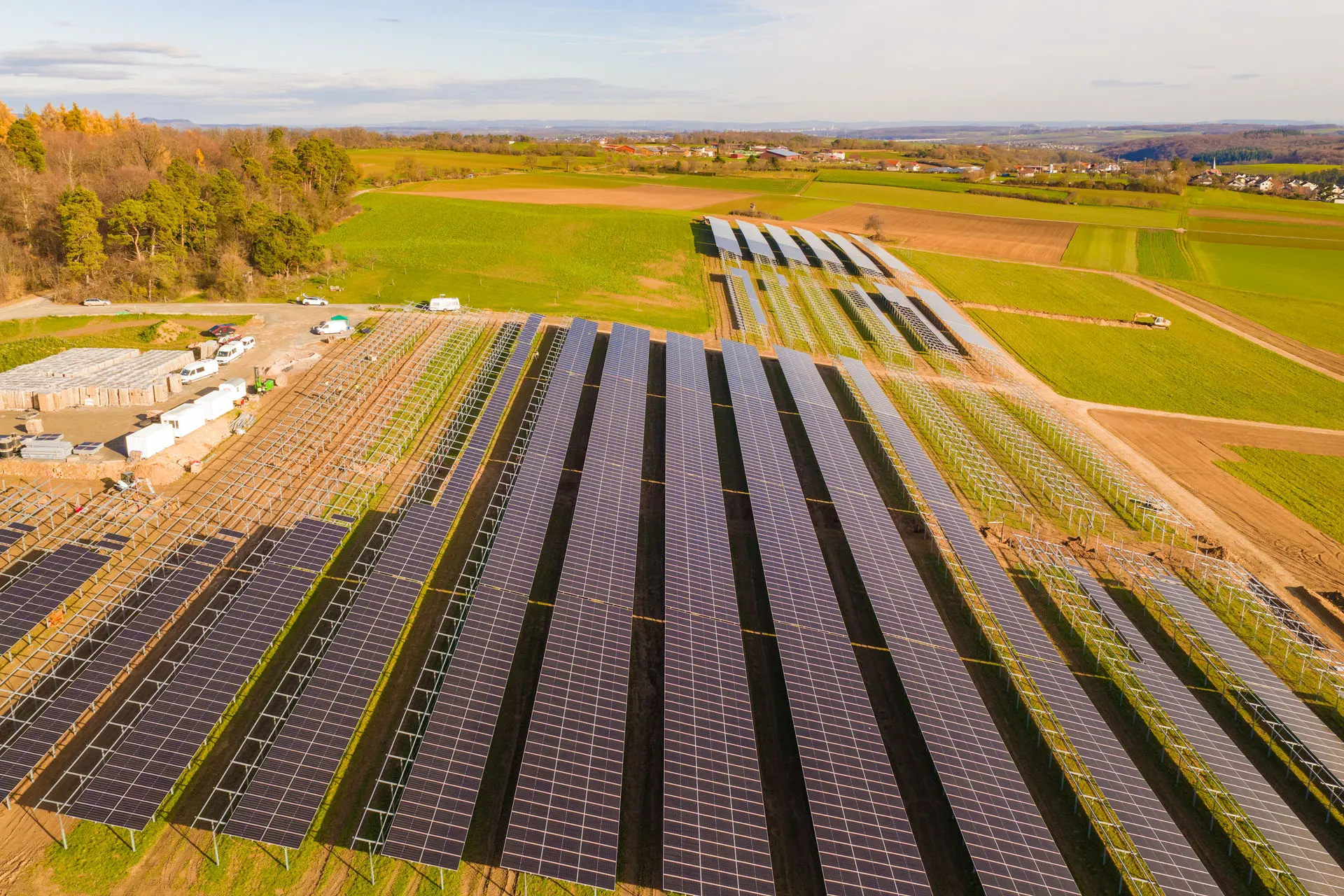 Große Photovoltaik-Freiflächenanlage im Bau; Reihen von Solarmodulen auf Metallgestellen in einer ländlichen Umgebung, daneben ein Baustellenlager mit Containern und Fahrzeugen.