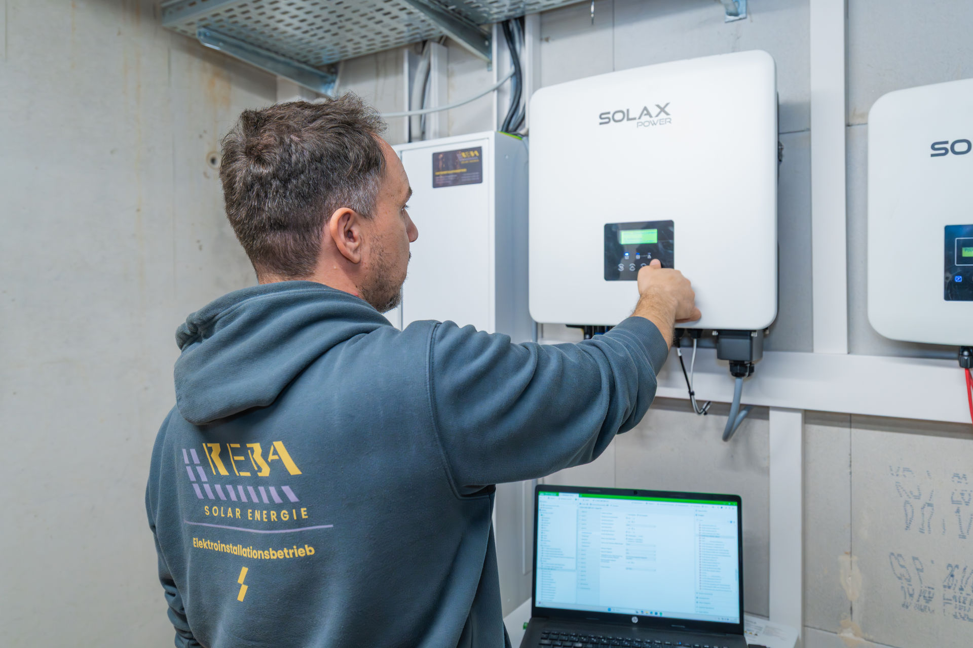 Ein Elektroniker von Reba Solar bedient das Display eines Solax Power Wechselrichters an einer Kellerwand, während ein Laptop im Vordergrund Daten anzeigt.