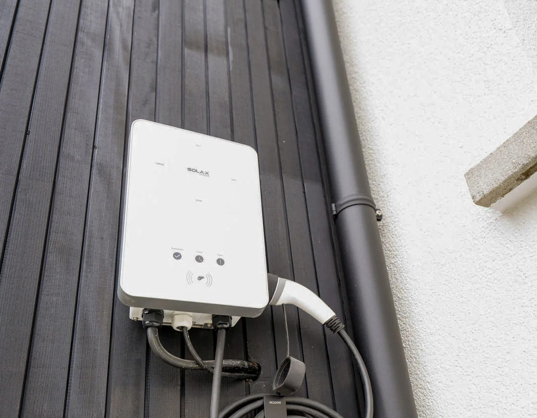 Eine an einer dunklen Holzfassade montierte SolaX Power Wallbox mit ordentlich aufgerolltem Ladekabel und eingestecktem Ladestecker.