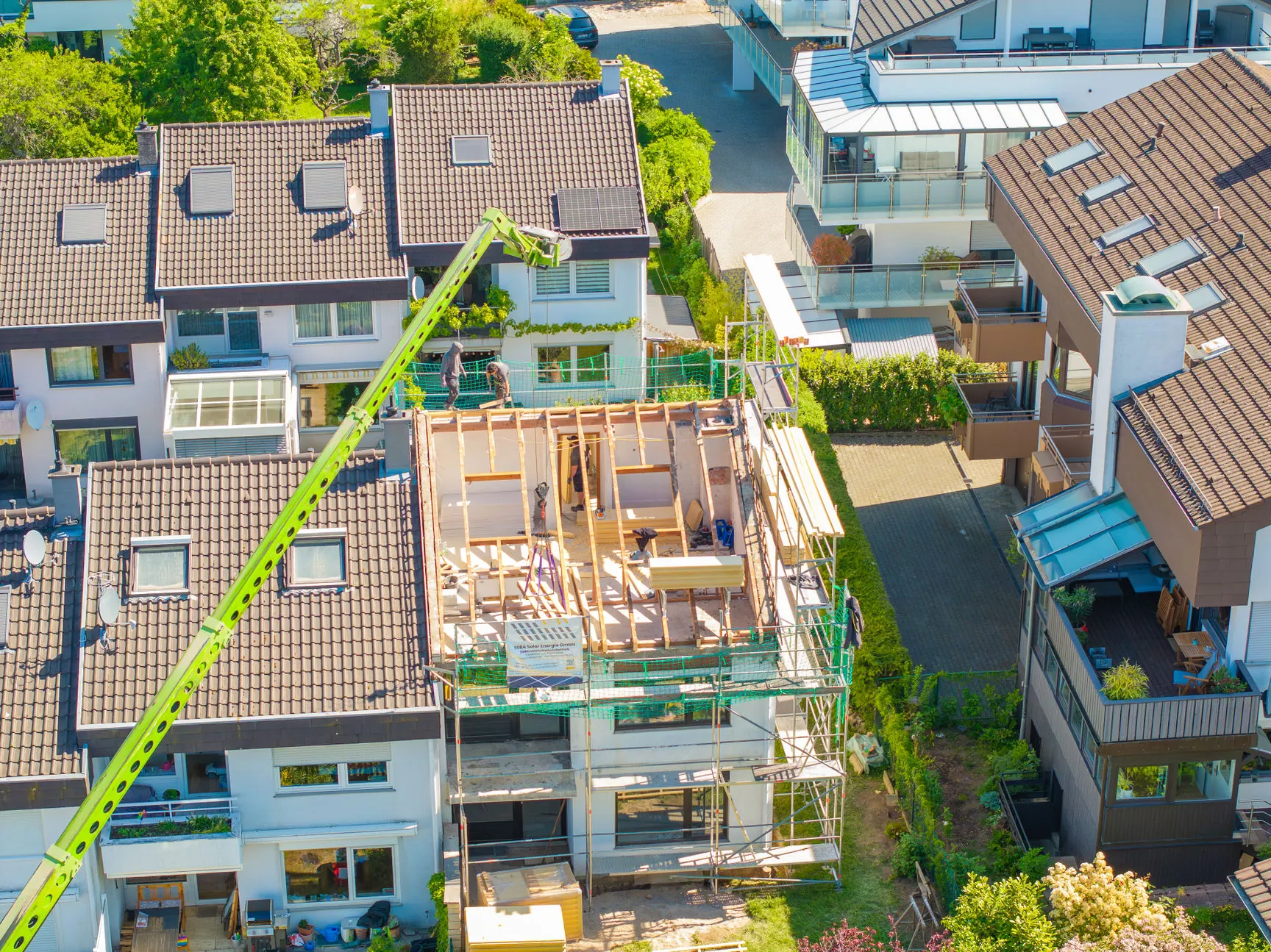Luftaufnahme einer Dachsanierung an einem Reihenhaus; ein Mobilkran hebt Baumaterial an den offenen Dachstuhl, der von einem Gerüst umgeben ist.