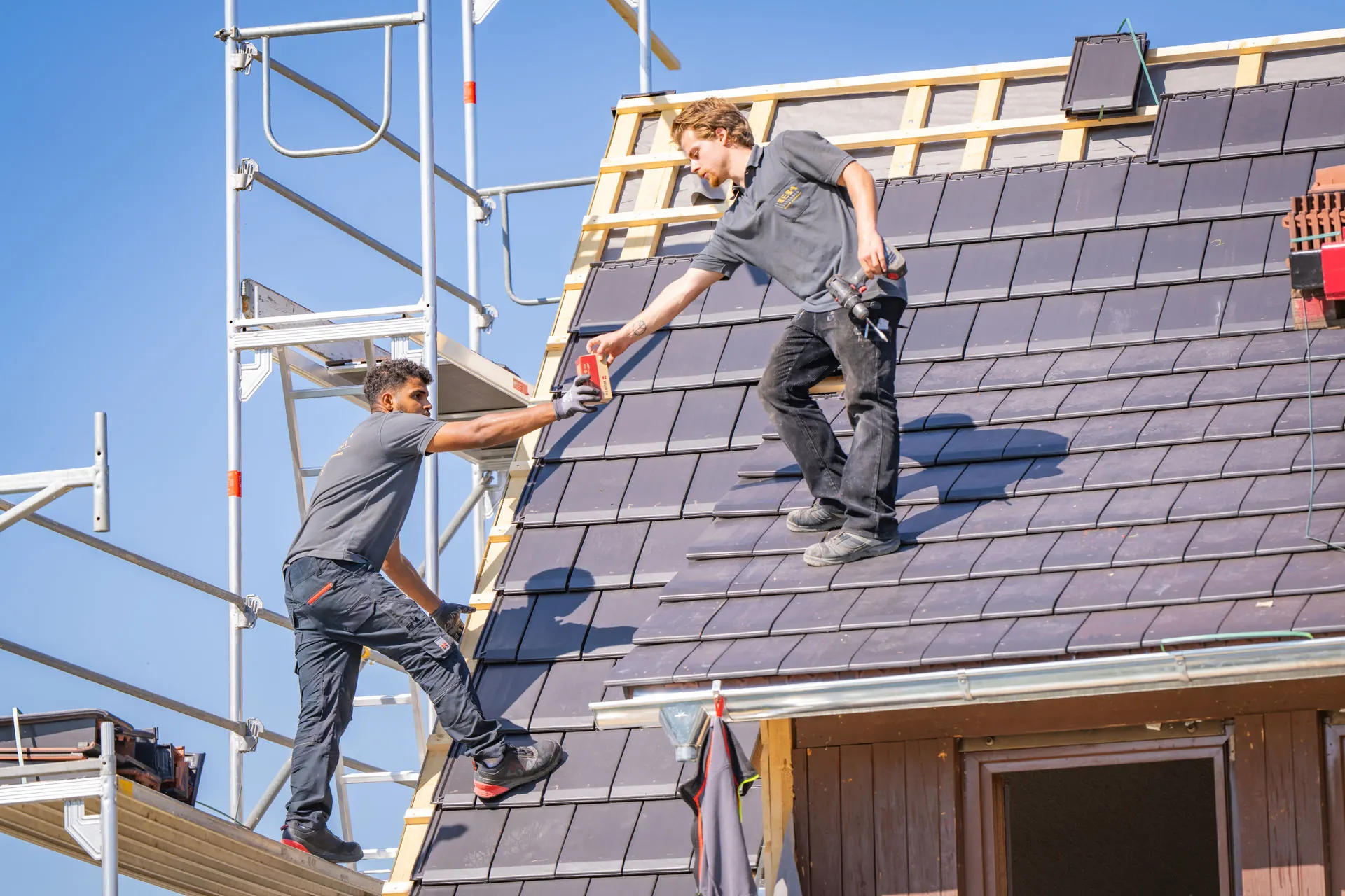 Zwei Dachdecker von Reba Solar bei der Eindeckung eines Steildachs mit anthrazitfarbenen Ziegeln; ein Mitarbeiter auf dem Gerüst reicht Material an seinen Kollegen auf der Dachfläche weiter.
