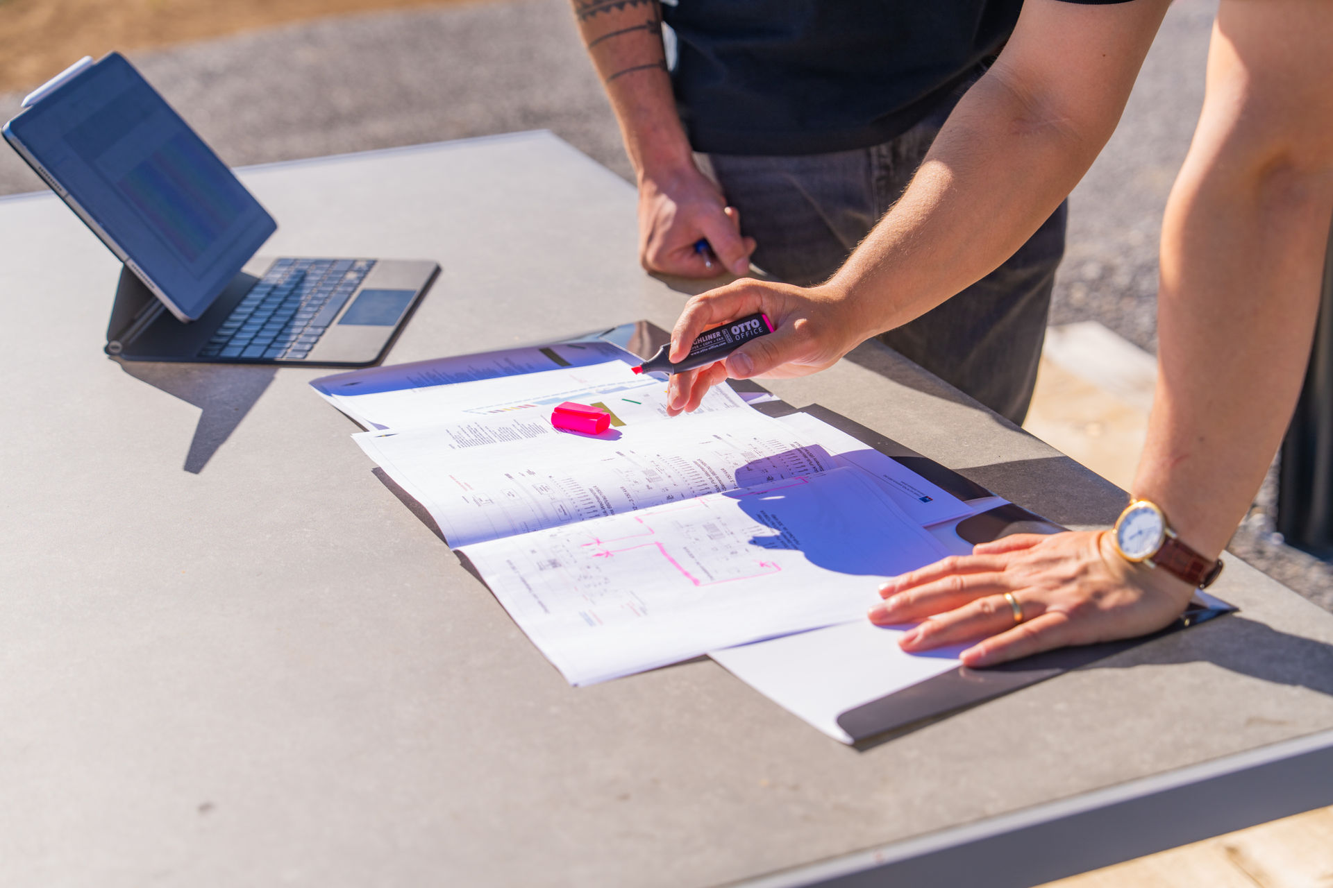 Projektplanung vor Ort: Hände von Fachkräften beim Analysieren von Dokumenten und Plänen auf einem Außentisch bei sonnigem Wetter.