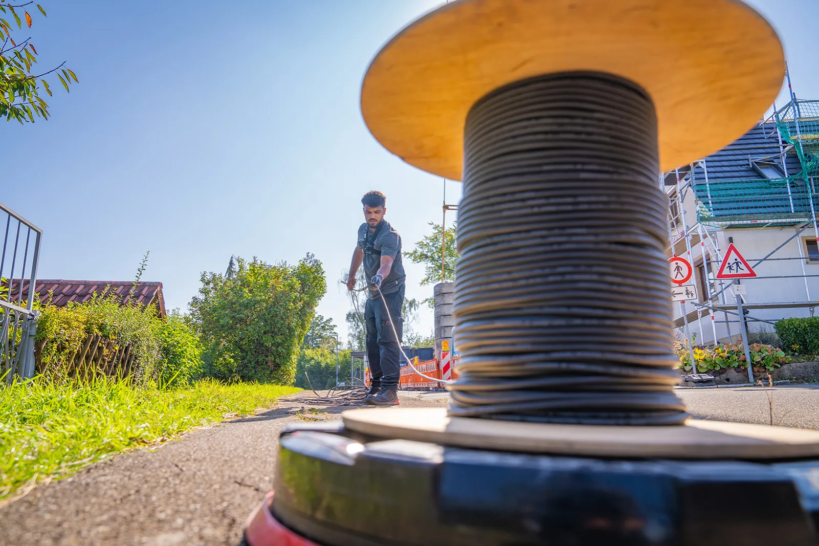 Ein Elektriker auf einer Baustelle im Freien, der ein schwarzes Kabel von einer großen Holzkabeltrommel abrollt; im Hintergrund ein Wohnhaus mit Gerüst.