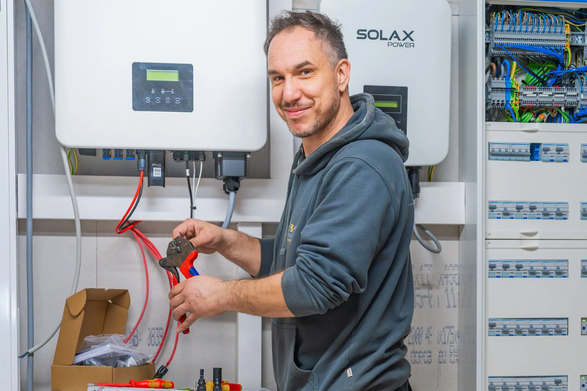 Ein Mann mittleren Alters mit Dreitagebart und einem dunkelgrauen Hoodie hält eine rot-blaue Crimpzange und ein rotes Kabel in den Händen. Er steht vor zwei weißen SolaX Power Wechselrichtern und einem offenen Stromverteilerkasten mit vielen Sicherungen und bunten Leitungen. Auf der Arbeitsfläche sind Werkzeuge und eine geöffnete Kartonschachtel zu sehen.