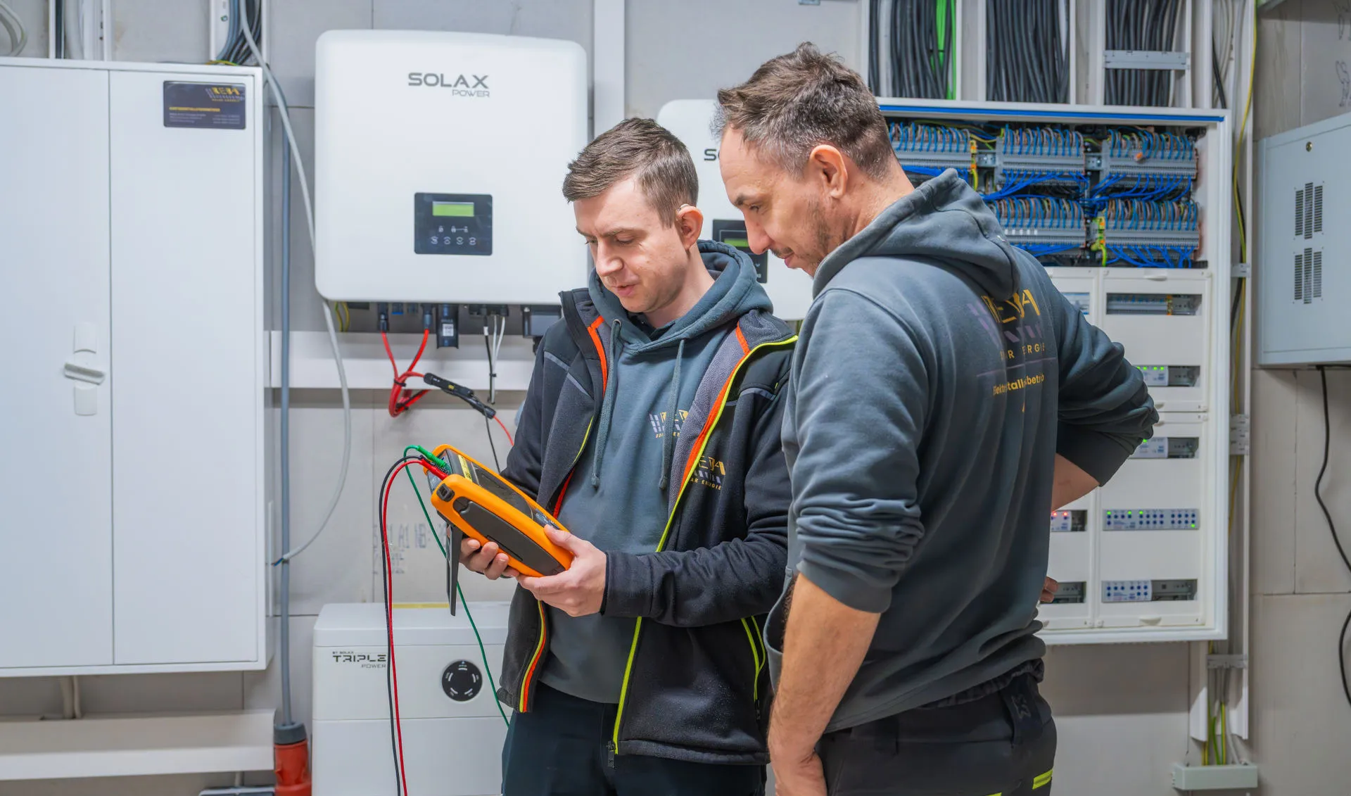 Zwei Elektriker prüfen eine Photovoltaik-Anlage mit einem orangefarbenen Messgerät; im Hintergrund sind SolaX-Wechselrichter und ein offener Schaltschrank an der Wand montiert.