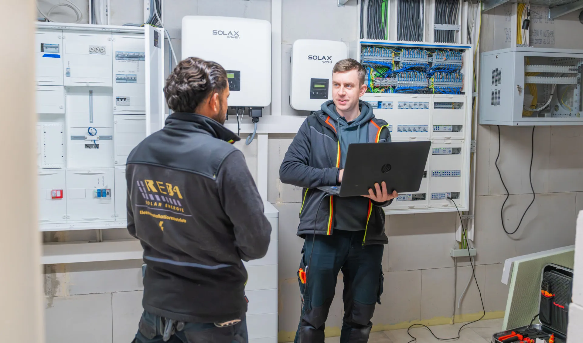 Fachberatung und digitale Inbetriebnahme einer Solaranlage mit Laptop vor einem offenen Sicherungskasten und Wechselrichtern.