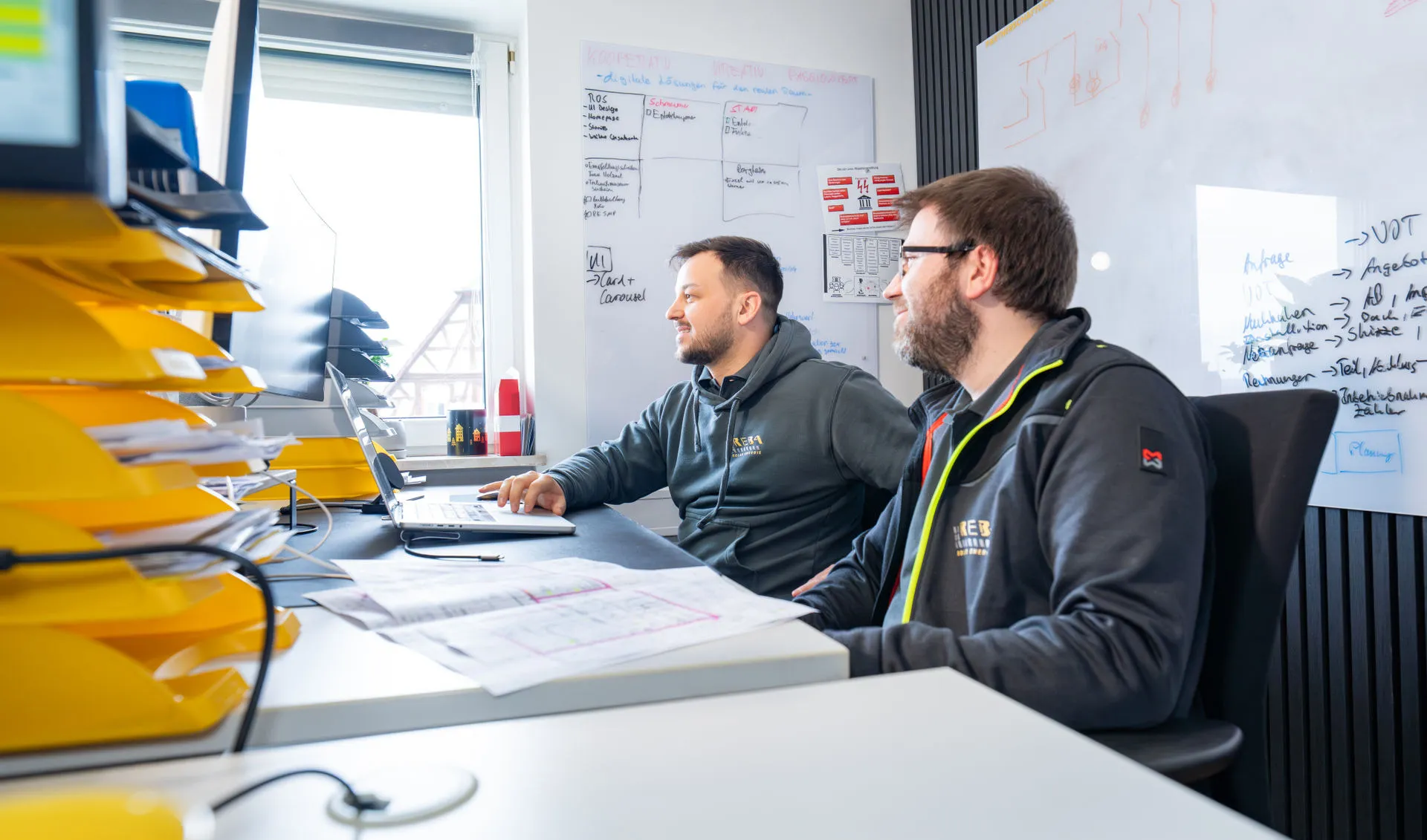 Zwei Männer in grauer Arbeitskleidung mit dem Logo „REBA Solar Energie“ sitzen konzentriert am Schreibtisch. Der linke Mann bedient einen Laptop, beide schauen nach links auf einen größeren Monitor. Vor ihnen liegen aufgeschlagene Schaltpläne. Links im Vordergrund steht ein Stapel gelber Ablagefächer. Der Hintergrund zeigt Whiteboards mit technischen Skizzen und Text.