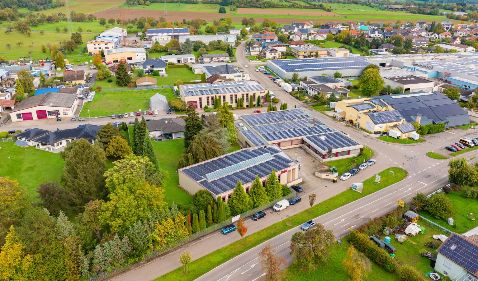Luftaufnahme eines Gewerbegebiets in ländlicher Umgebung, wobei der Fokus auf mehreren großen Hallendächern liegt, die flächendeckend mit Photovoltaik-Anlagen ausgestattet sind. Im Hintergrund ist ein Dorf und landwirtschaftliche Flächen zu sehen.