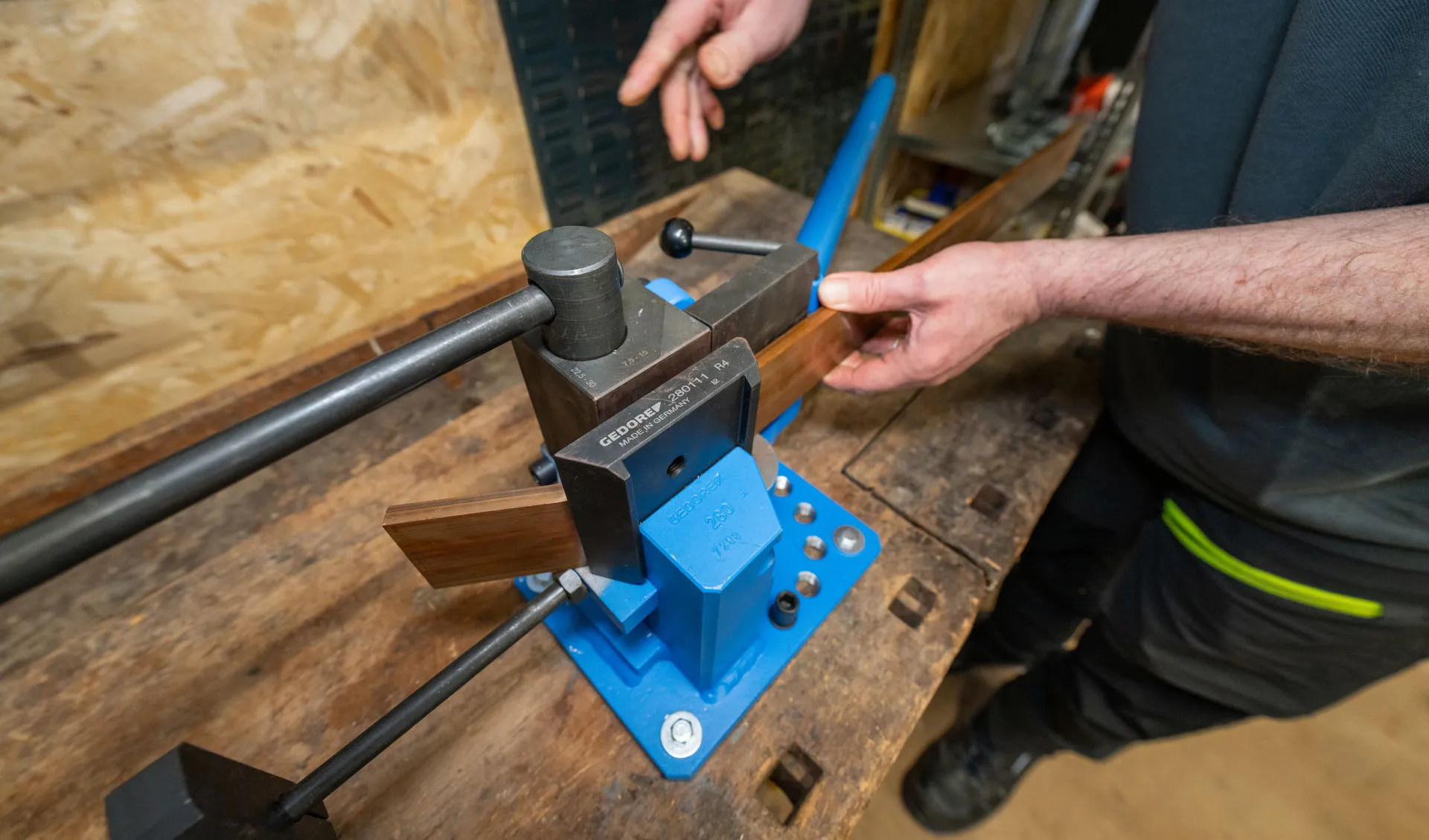Nahaufnahme einer handbetriebenen Metallbiegemaschine mit blauer Basis und schwarzen Griffen auf einer abgenutzten Holzwerkbank. Ein Mann in Arbeitskleidung führt mit beiden Händen ein flaches Kupferband durch die Biegewerkzeuge der Maschine. Die Gravur "GEDORE" und "MADE IN GERMANY" ist auf dem Werkzeugkopf deutlich lesbar. Im Hintergrund ist eine OSB-Holzwand und eine andere Person zu sehen.
