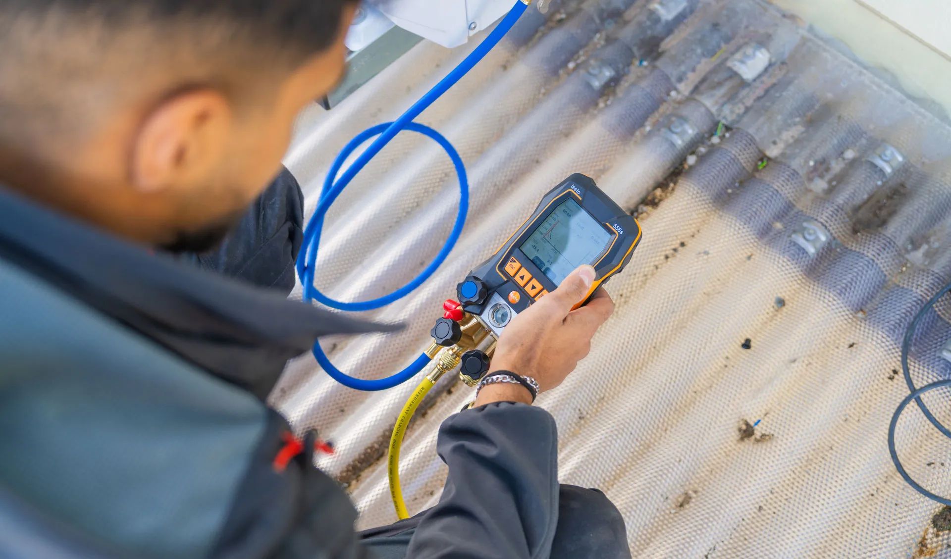 Blick über die Schulter eines Technikers, der ein digitales Messgerät (Monteurhilfe) der Marke Testo in den Händen hält. Das Gerät zeigt Grafiken und Messwerte auf einem beleuchteten Display an. Gelbe und blaue Serviceschläuche sind an das Gerät angeschlossen. Der Techniker trägt dunkle Arbeitskleidung und befindet sich auf einem hellen, leicht verschmutzten Wellplatten-Dach.