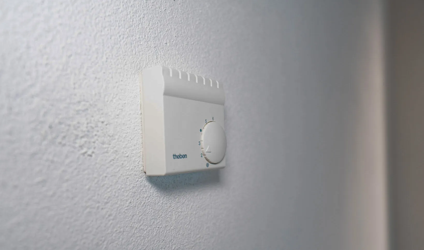 Ein analoges Wandthermostat der Marke Theben mit einem runden Drehregler zur Temperatursteuerung, montiert auf einer weiß verputzten Wand.