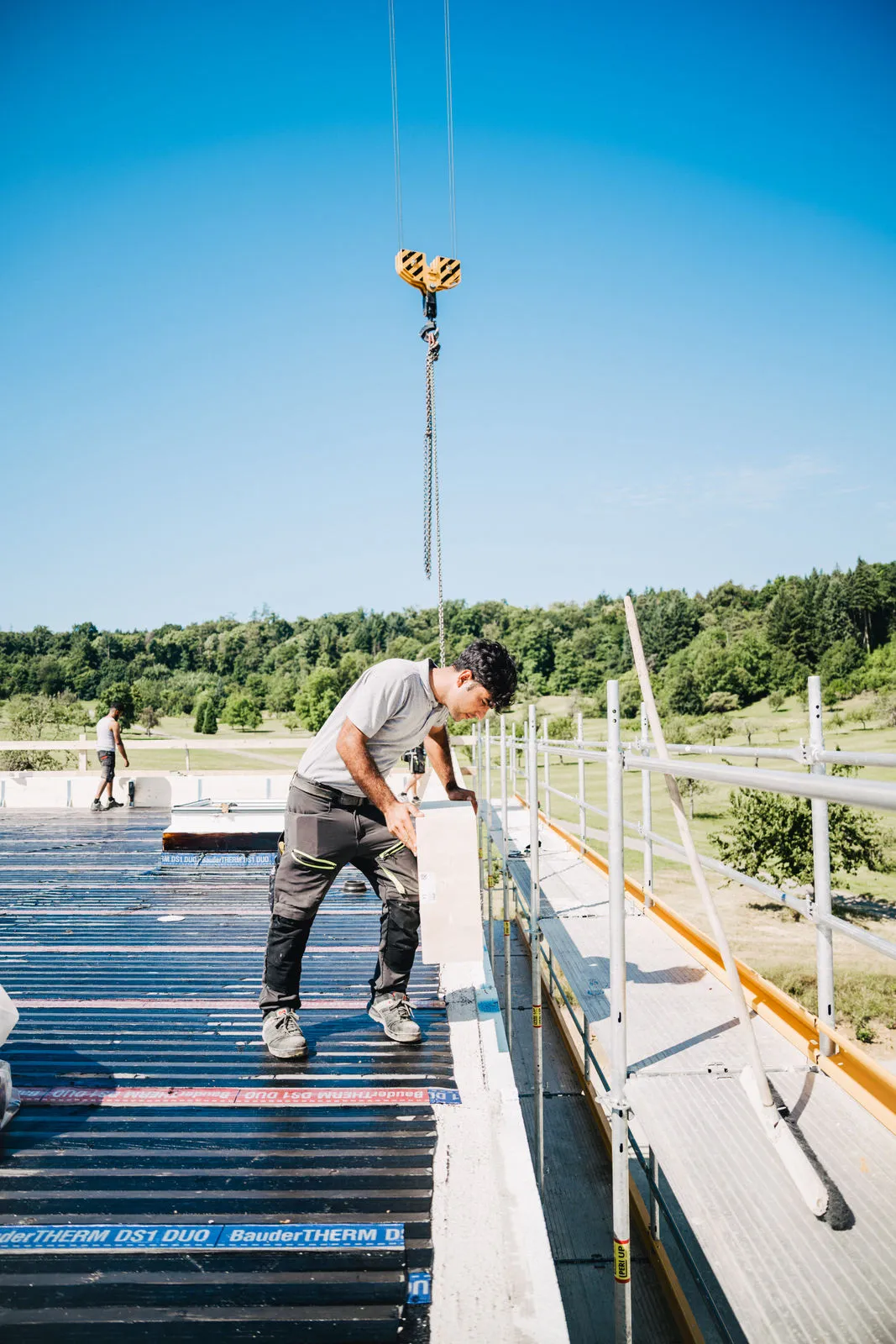 Fachgerechte Installation der Dachabdichtung mit BauderTHERM-Bahnen und Materialtransport per Kran auf einer Flachdach-Baustelle.