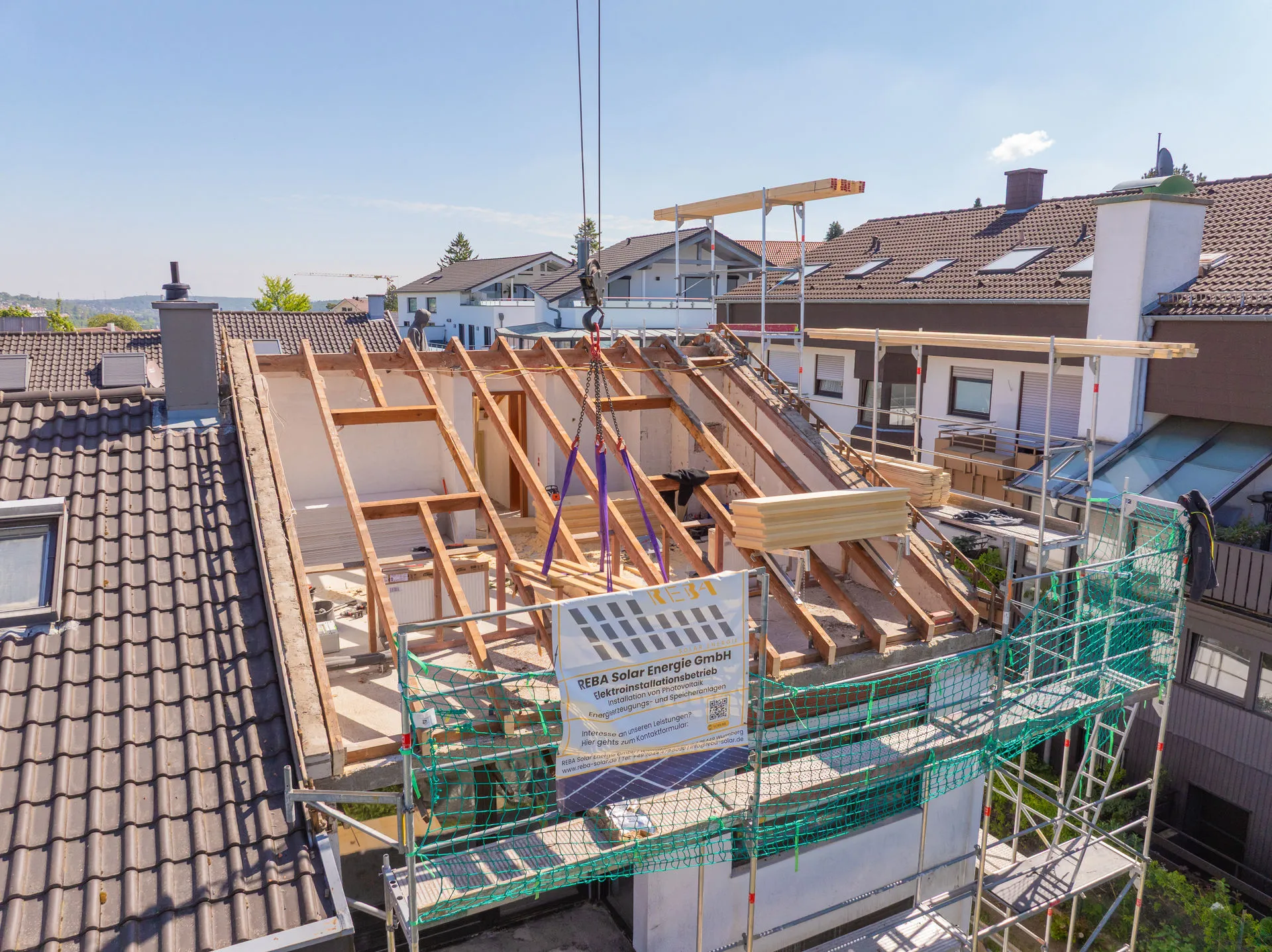 Luftaufnahme eines im Bau befindlichen Dachstuhls aus Holz. Ein Kran hebt Baumaterialien an, und ein Bauschild der Firma REBA Solar Energie GmbH ist am Gerüst angebracht.